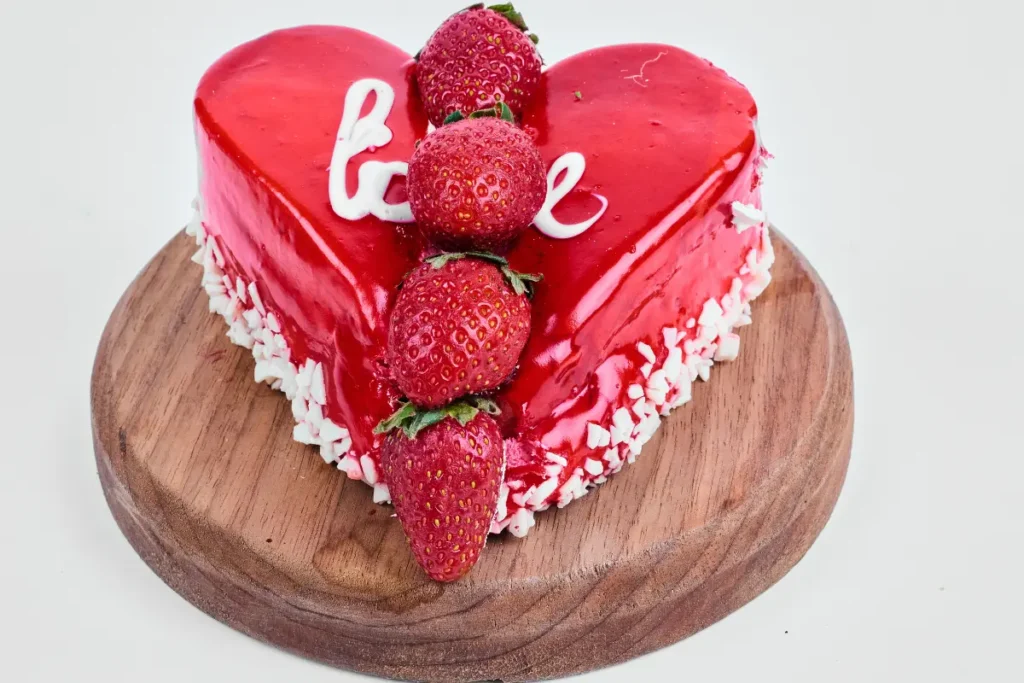 Elegant pink heart-shaped cake adorned with edible flowers and pearls on a white cake stand, symbolizing love and celebration.