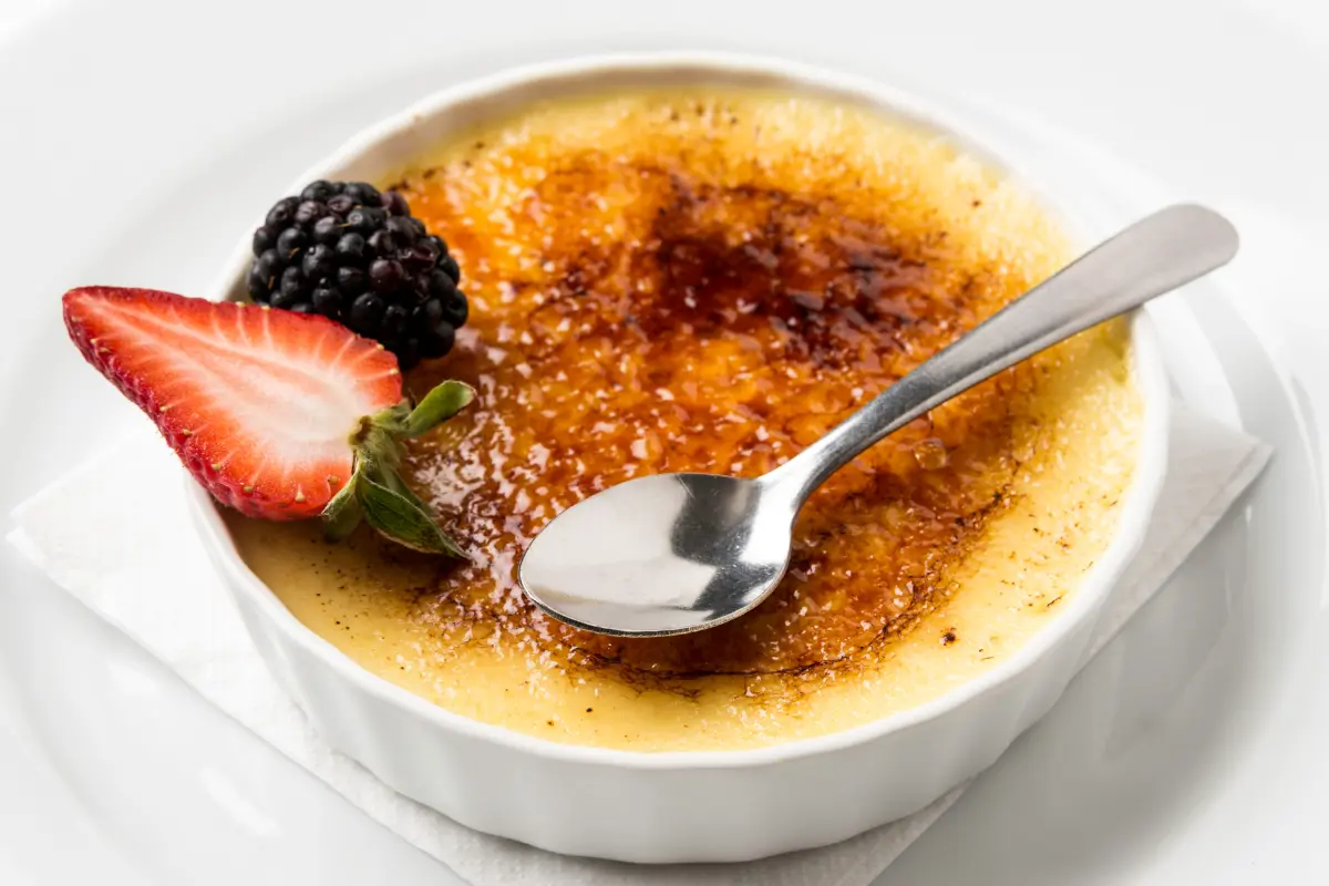 Golden-brown crab brulee in white ramekin with fresh dill and lemon garnish on a dark wooden table.